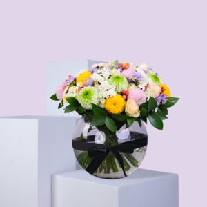 A colorful bouquet of flowers in a round glass vase, tied with a black ribbon. The bouquet includes chrysanthemums, roses, and other vibrant flowers.