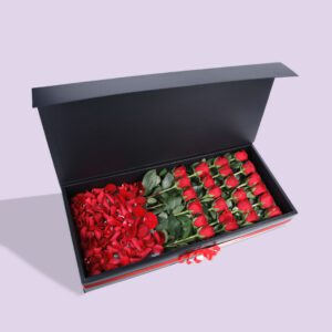 A black gift box open to reveal a compartment of red roses and another compartment of red rose petals.